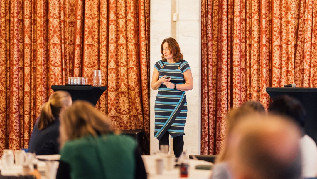 En kvinne i en blåstripet kjole står og snakker foran et publikum på en konferanse, med røde mønstrede gardiner i bakgrunnen.