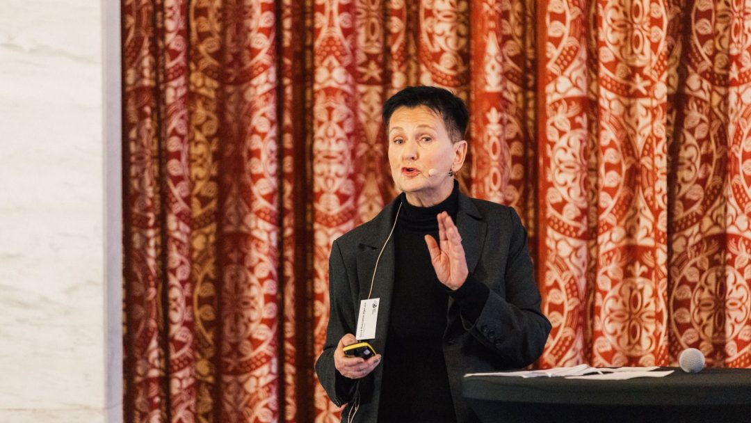 En person i svart antrekk snakker inn i en mikrofon mens han gestikulerer, og står foran røde mønstrede gardiner ved siden av et bord med papirer og en mikrofon.