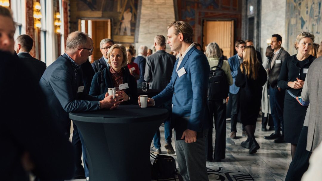 Folk i forretningsantrekk står og prater rundt cocktailbord i et stort, elegant rom med veggmalerier på veggene.
