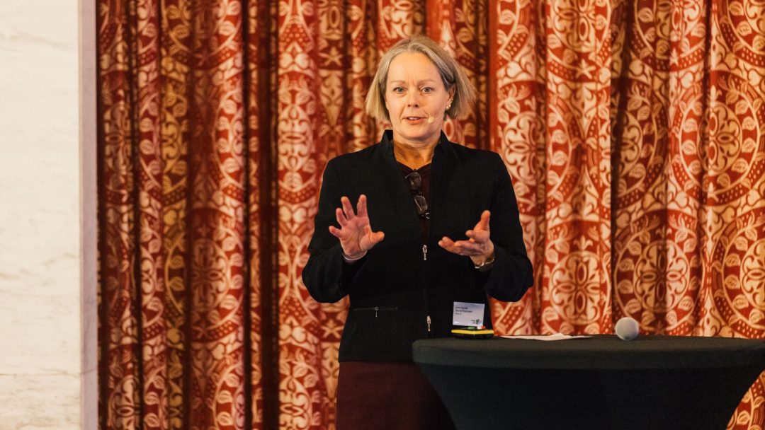 En person står og snakker foran et rødt og gullmønstret gardin og gestikulerer med hendene. En mikrofon er på et bord i nærheten.