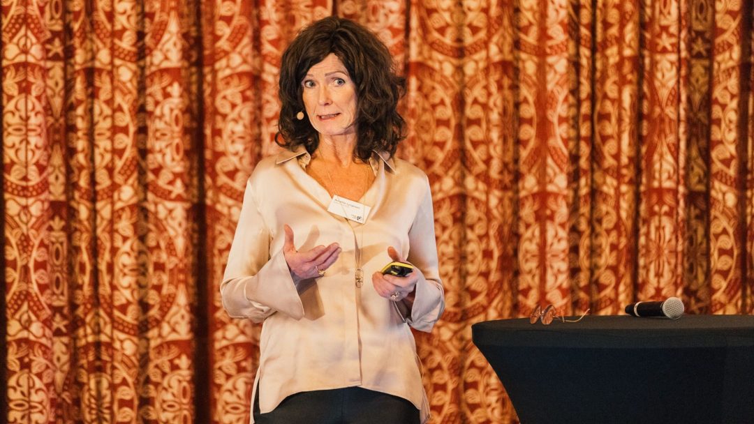 En kvinne med mikrofon gir en presentasjon foran utsmykkede røde gardiner og holder en klikker. En mikrofon ligger på et lite dekket bord ved siden av henne.