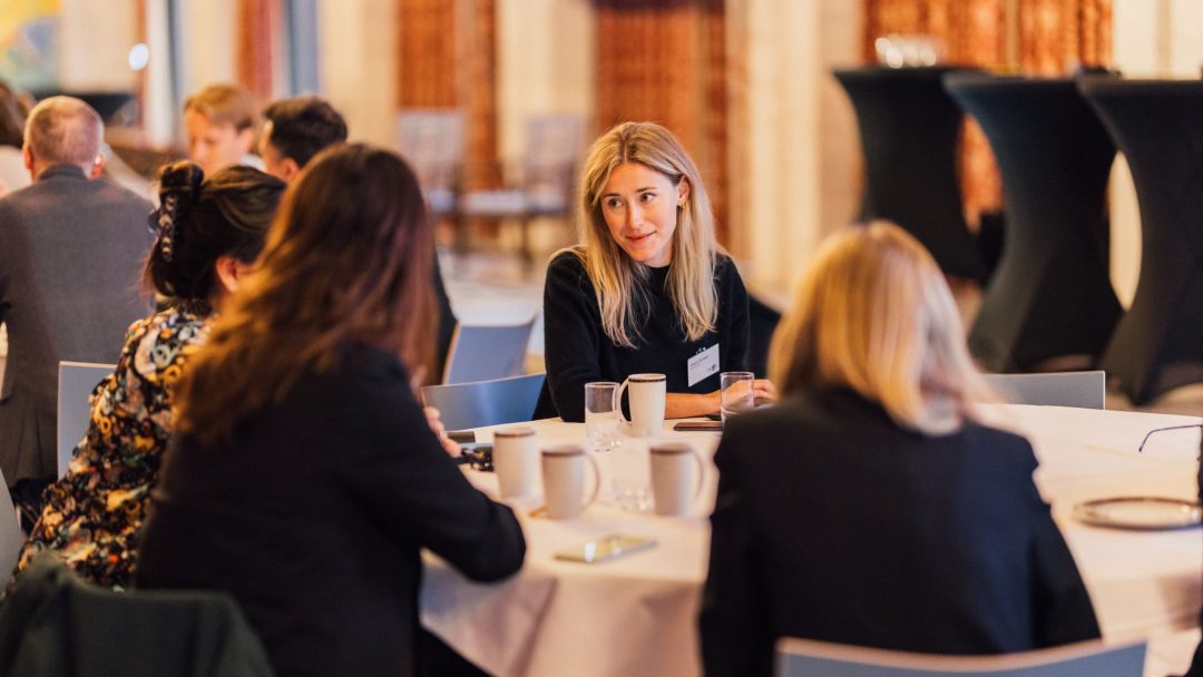 En gruppe mennesker sitter rundt et bord med kaffekopper, engasjert i samtale i et konferanserom.