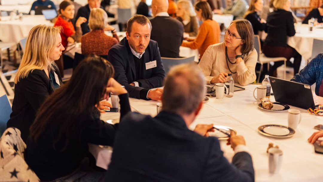 En gruppe mennesker engasjerte seg i diskusjon rundt et bord på et innendørs arrangement.