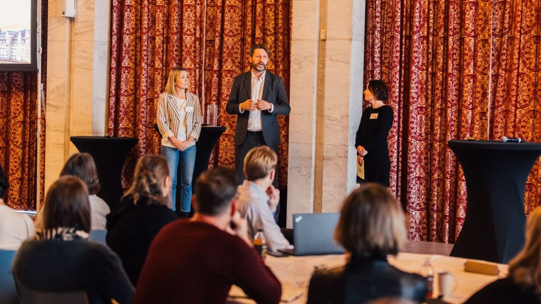 Tre personer står foran et publikum og snakker på en konferanse eller et arrangement. En mann i sentrum snakker mens to kvinner står på hver side. Rommet har rødmønstrede gardiner.