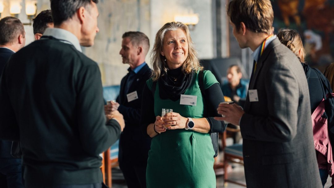 En gruppe mennesker i forretningsantrekk er samlet i samtale på et innendørs arrangement. En kvinne i en grønn kjole smiler og holder en drink blant dem.