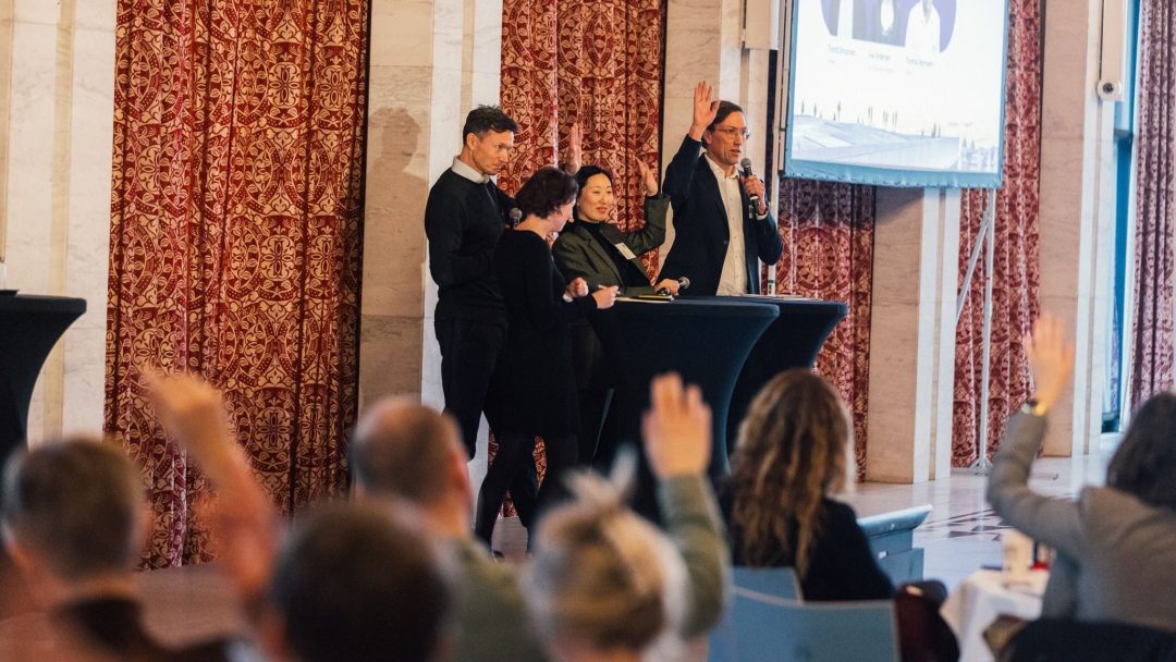En gruppe på fire personer står bak podier, en mann snakker i en mikrofon. Publikumsmedlemmer med løftede hender er delvis synlige i forgrunnen.