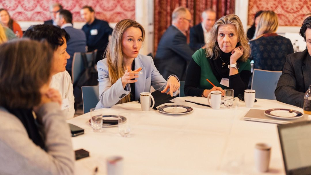 En gruppe mennesker sitter rundt et bord i diskusjon, med kaffekopper og tallerkener foran seg, i et rom med rødmønstrede vegger og gardiner.
