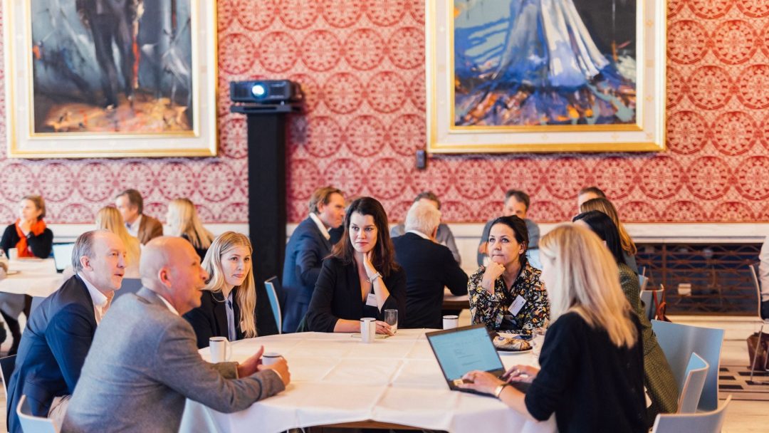 Folk satt rundt bordene i et rom med utsmykkede røde tapeter og store innrammede malerier, engasjert i samtale, med en bærbar datamaskin åpen på ett bord.