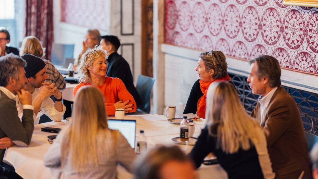 En gruppe mennesker som sitter rundt et bord, engasjert i samtale, i et rom med rødmønstrede vegger og utsmykket med et innrammet maleri.