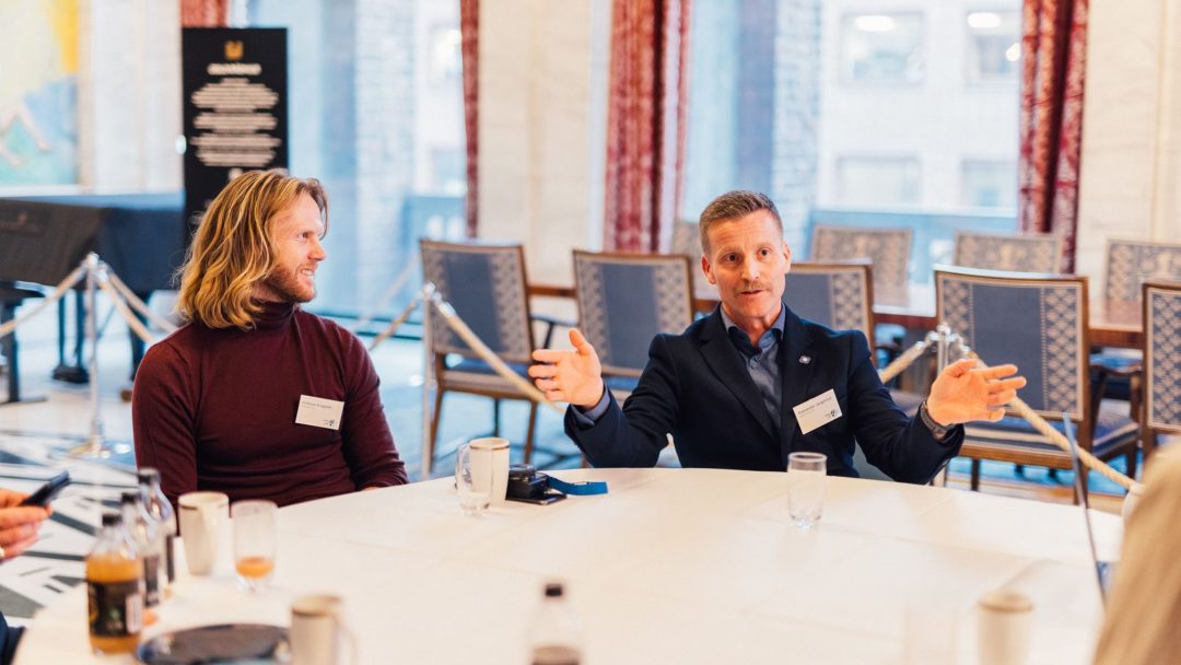 En gruppe mennesker sitter rundt et rundt bord i et møterom, engasjert i samtale. En mann gestikulerer med hendene mens han snakker.