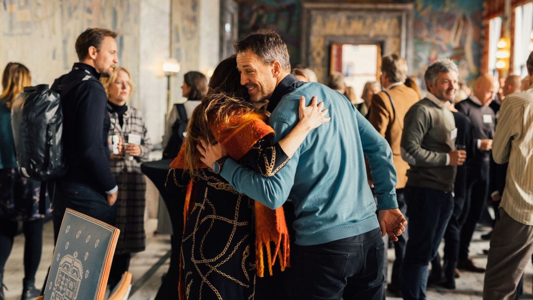 En gruppe mennesker som sosialiserer seg på et innendørs arrangement; to individer omfavner seg i forgrunnen.