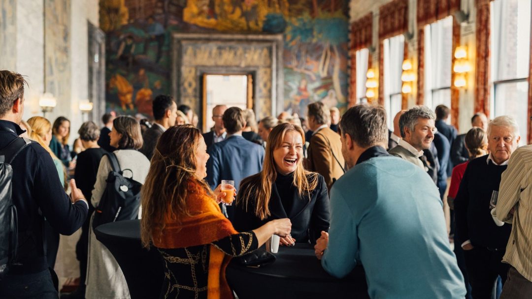 Folk mingler og snakker på en sosial sammenkomst i et stort, utsmykket rom med veggmalerier og store vinduer.