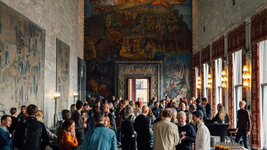 En stor gruppe mennesker blander seg i et utsmykket rom med steinvegger, veggmalerier og høye vinduer. Det er et bord med forfriskninger i forgrunnen.