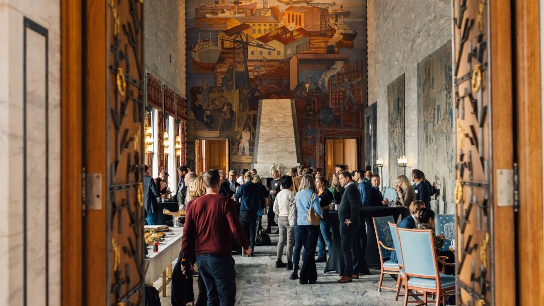 Folk samles i et stort, utsmykket rom med veggmalerier på veggene og et åpent buffébord.