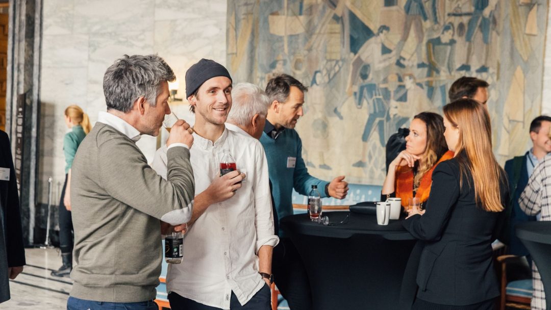 Folk som sosialiserer og drikker på et nettverksarrangement i en formell setting med et stort veggmaleri i bakgrunnen.
