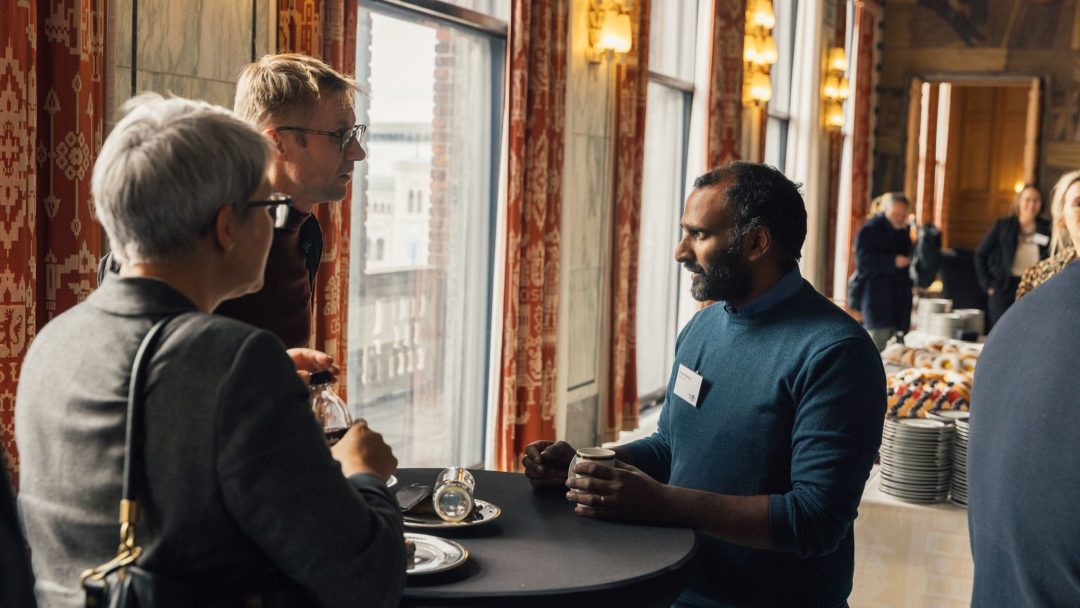 Tre personer står rundt et høyt bord og snakker i et varmt opplyst rom. Det er mer folk og forfriskninger i bakgrunnen.