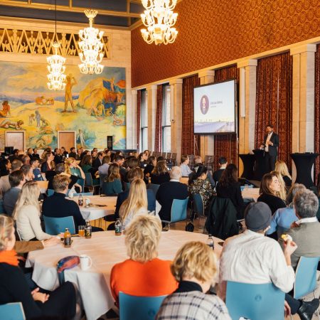 En foredragsholder henvender seg til et publikum som sitter ved bordene i et stort, utsmykket rom med lysekroner og et veggmaleri.