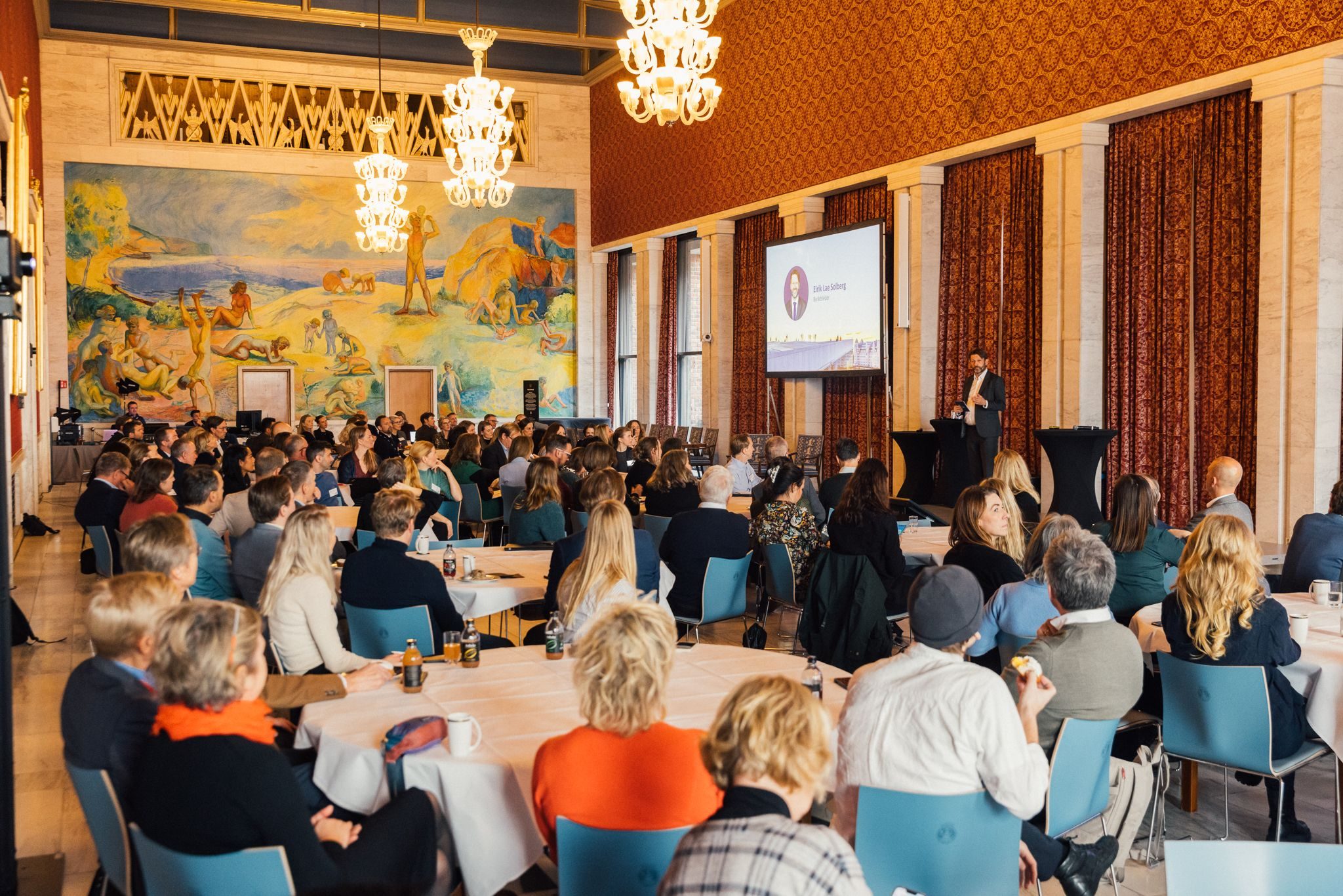 En foredragsholder henvender seg til et publikum som sitter ved bordene i et stort, utsmykket rom med lysekroner og et veggmaleri.