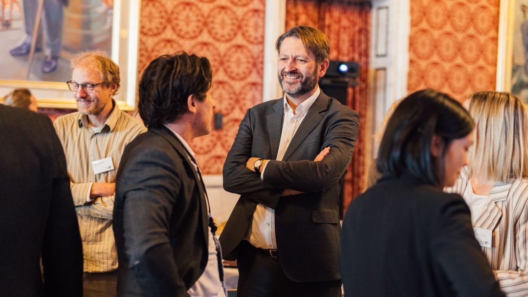 En gruppe mennesker er i samtale på et innendørs arrangement. En person i dress står med armene i kors, mens andre møter hverandre i diskusjon. Rommet har utsmykkede tapeter.