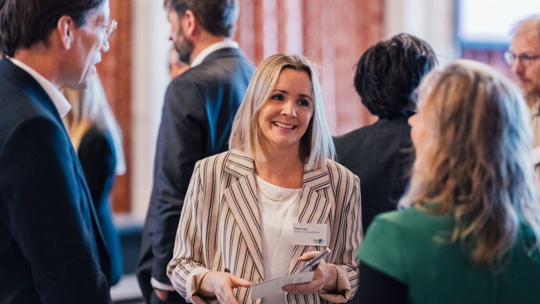 En kvinne i en stripet blazer med navnelapp snakker med to personer i en gruppe. Andre individer er synlige i bakgrunnen.