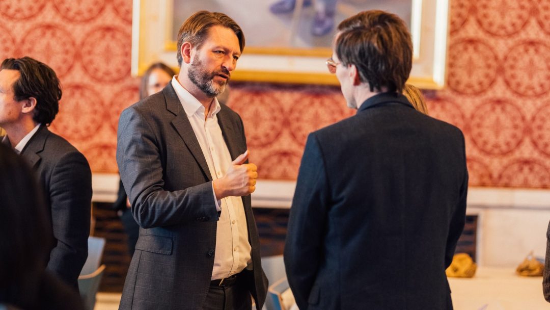 Flere personer i forretningsantrekk er engasjert i samtale i et formelt rom med en rød mønstret vegg og et stort innrammet bilde.