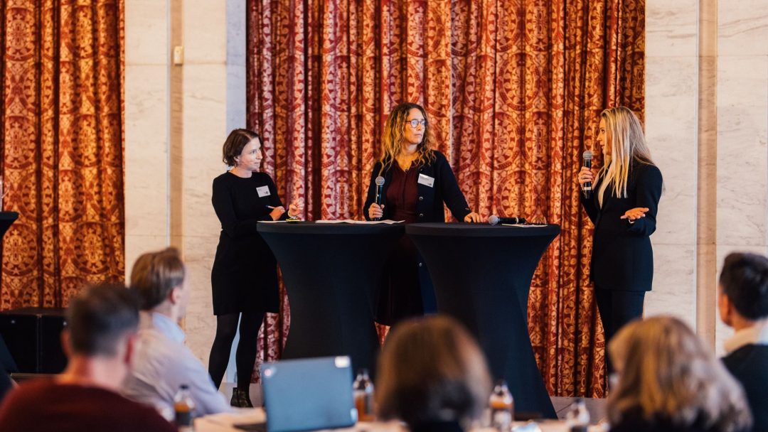 Tre kvinner står bak bord og snakker i mikrofoner på en konferanse, vendt mot et publikum som sitter med bærbare datamaskiner og drinker.