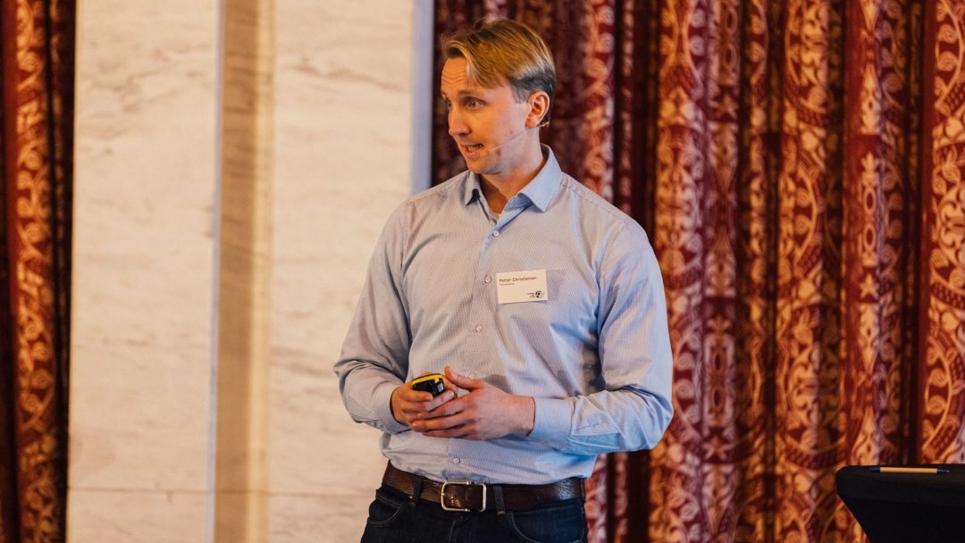 Person i blå skjorte som snakker på et arrangement, står foran utsmykkede røde gardiner og holder en liten enhet.