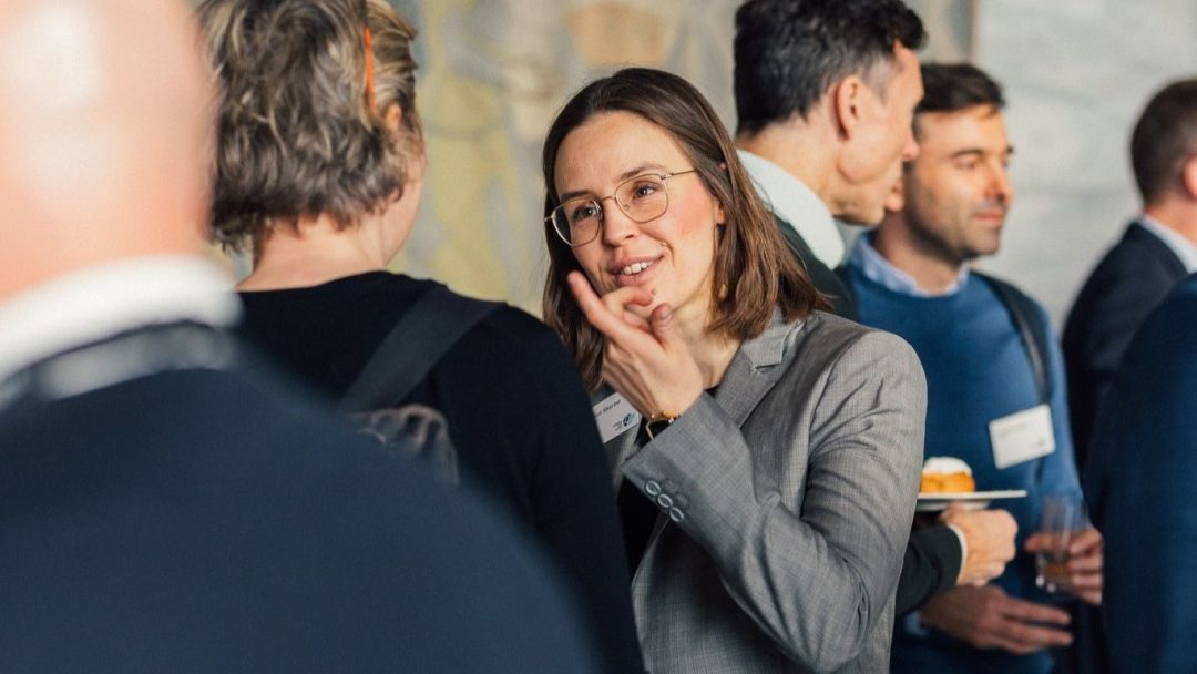 En kvinne i grå dress og briller deltar i samtale på et nettverksarrangement. Folk i forretningsantrekk er i bakgrunnen.
