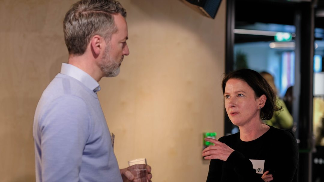 To personer snakker innendørs. Mannen holder et glass vann og har på seg en lyseblå genser. Kvinnen gestikulerer med hånden og har på seg en svart topp.