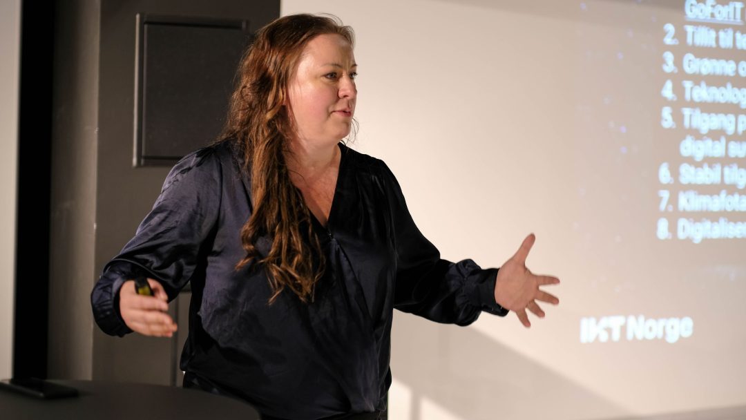 En person med langt hår og svart skjorte snakker ved et podium og gestikulerer med hendene. En presentasjon med tekst er delvis synlig på en skjerm bak.