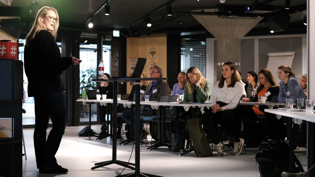 Person som står og snakker til et sittende publikum i et moderne konferanserom. Bord og bærbare datamaskiner er synlige, med en oppmerksom gruppe som ser på presentasjonen.