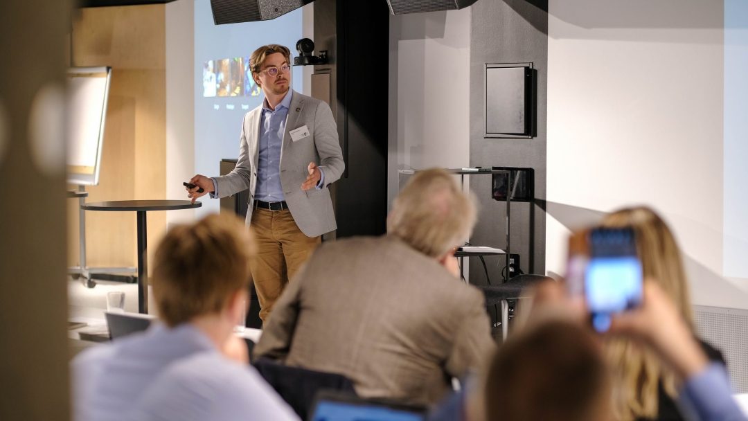 En mann i en grå blazer presenterer på en konferanse og snakker til et publikum. Noen deltakere tar notater på bærbare datamaskiner eller bruker smarttelefoner.