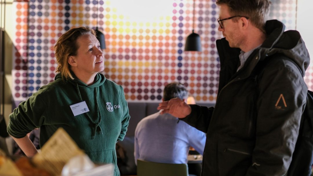 To personer snakket innendørs, med en fargerik vegg med prikker i bakgrunnen. En person har på seg en grønn hettegenser, den andre en mørk jakke og briller.