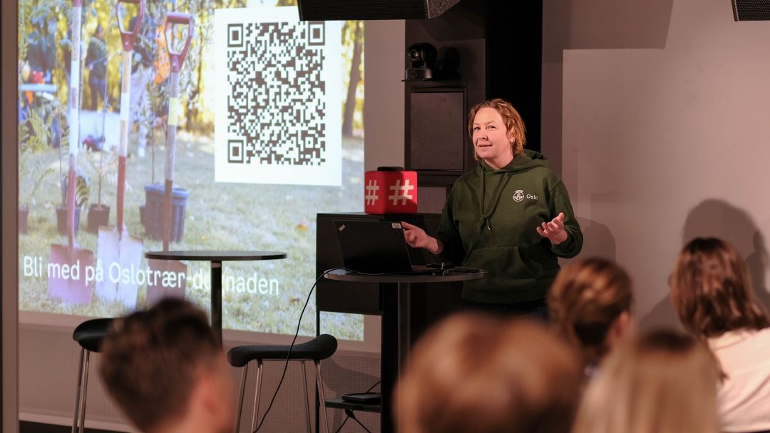 En person snakker på en presentasjon foran et publikum. En skjerm viser en QR-kode og tekst på et annet språk.