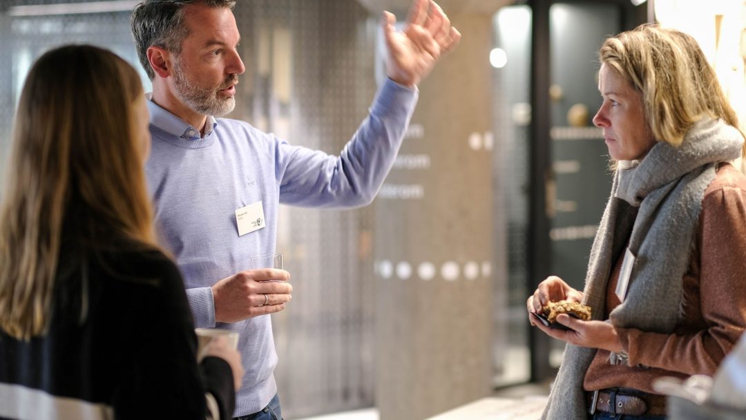 Tre personer har en samtale innendørs, med en mann som gestikulerer mens han holder en drink og to kvinner som lytter, en holder en tallerken med mat.