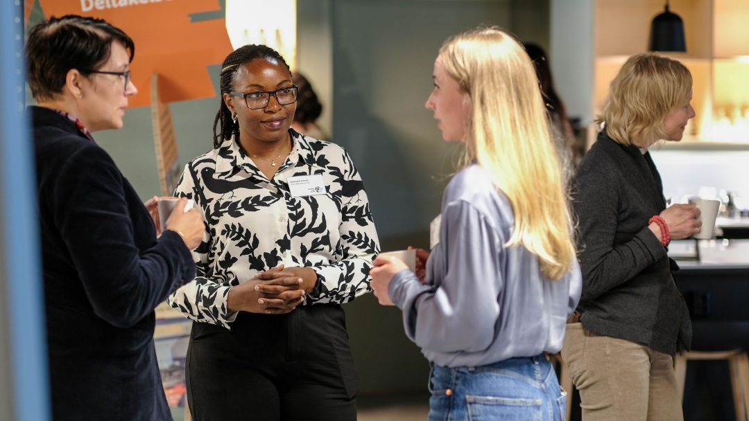 Fire personer er engasjert i samtale i en innendørs setting, med et skilt med Deltakelse i bakgrunnen.