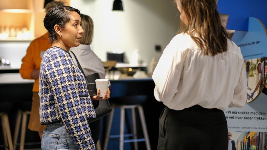 To kvinner står i samtale innendørs; man holder en kopp. De er i nærheten av en disk med krakker, og et stativ med en plakat er delvis synlig.