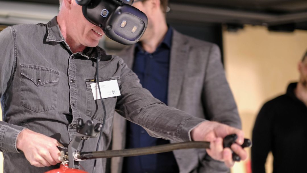 Person som har på seg et VR-hodesett, holder en brannslukningsslange som om han bruker den, med to personer i bakgrunnen som observerer.