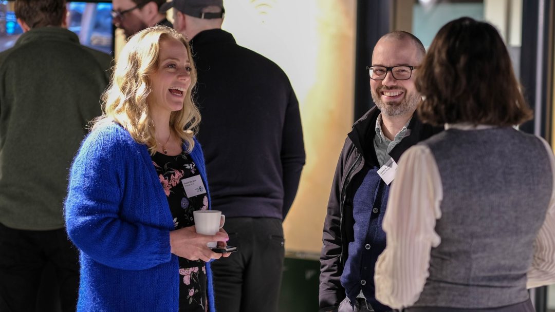 Tre personer var i samtale på et innendørs arrangement. En kvinne som holder en kopp smiler, mens en mann med briller lytter. Andre deltakere er i bakgrunnen.