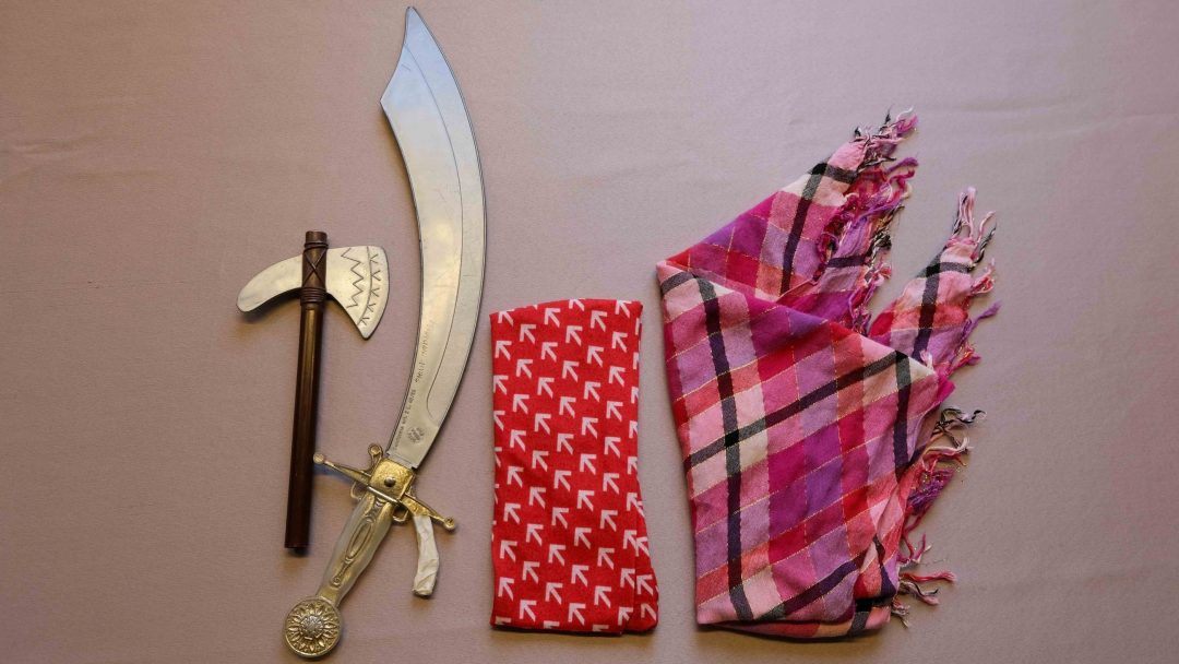 Et buet sverd, en liten øks med treskaft og et rødt og hvitt mønstret tekstil lå på en grå overflate. I nærheten, perfekt for enhver halloweenkostyme, hviler et rosa rutete skjerf.