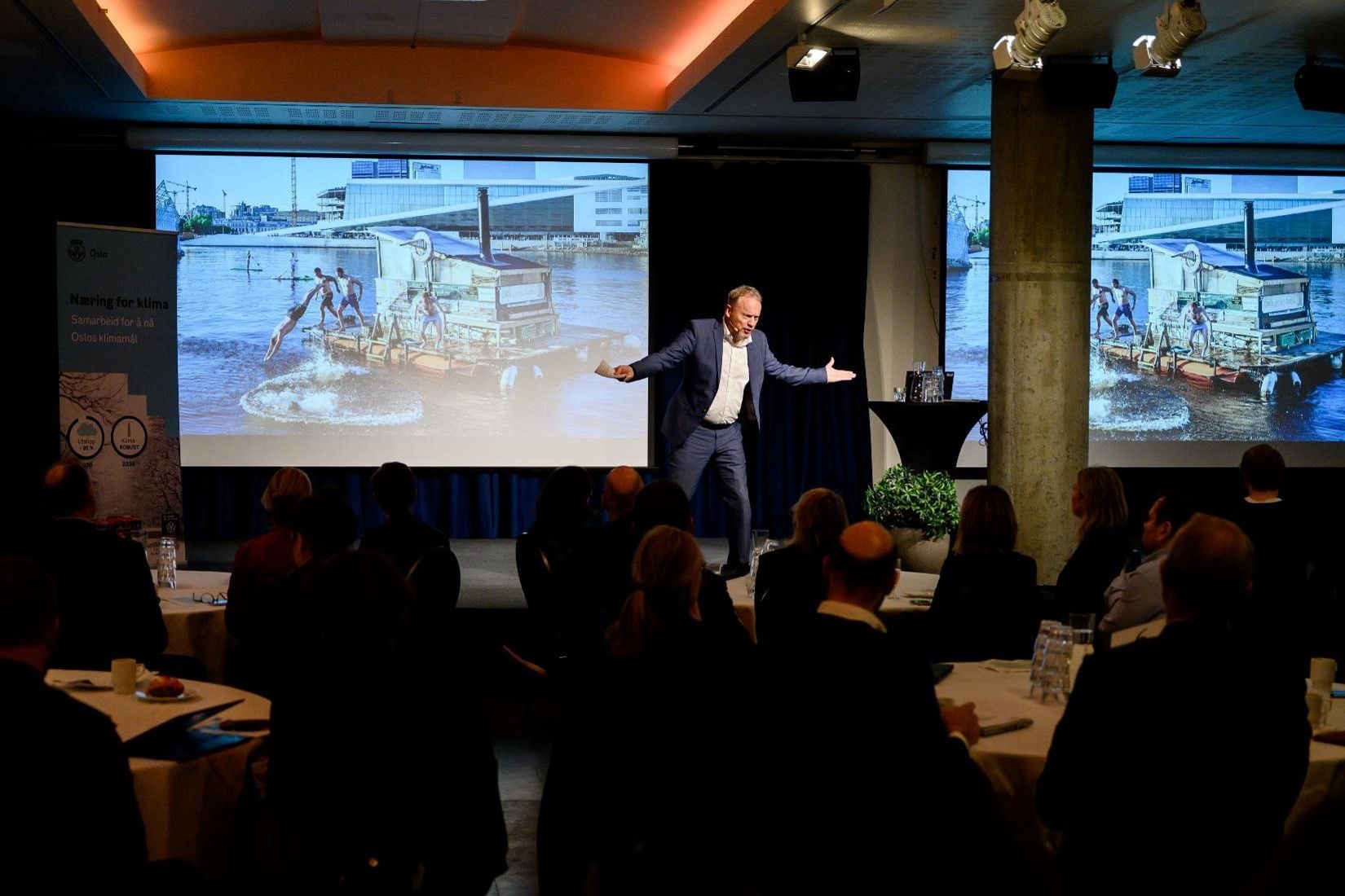 En mann gestikulerer på scenen i et konferanserom, med en stor projeksjon av et bilde som viser to personer som sykler på vannet. Publikumsmedlemmer sitter rundt runde bord.
