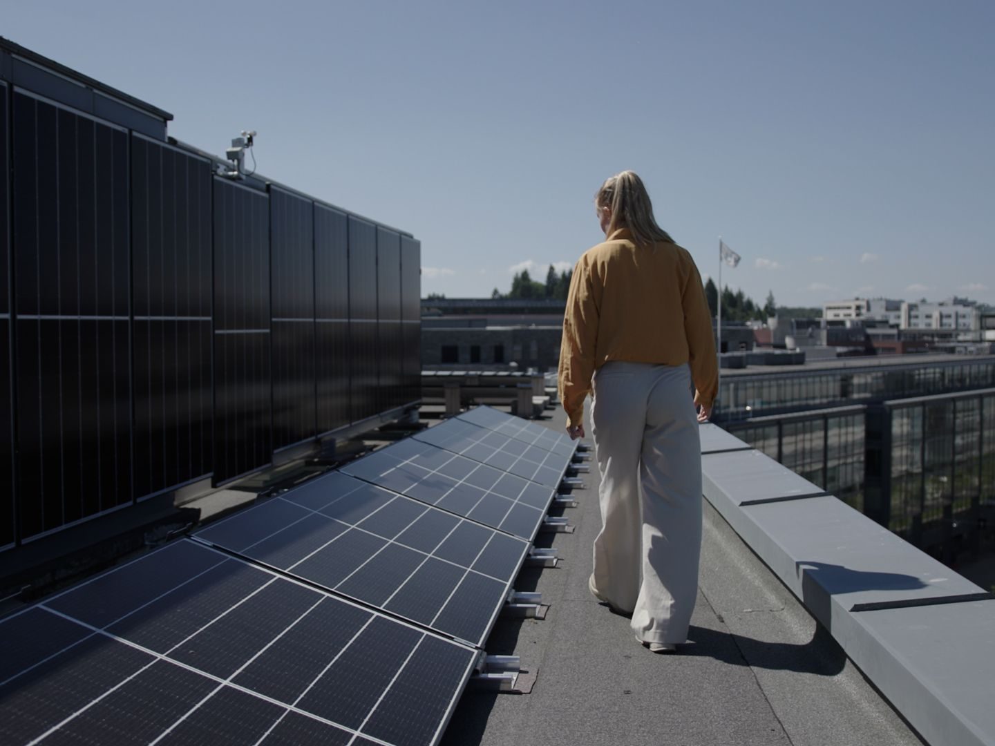 Midt i et bakteppe av bybygninger og en klar blå himmel, rusler en person over taket og navigerer mellom rader med solcellepaneler. Kledd i en brun jakke og lyse bukser, legemliggjør de prinsippene skissert i klimabudsjettet – fremme bærekraftig liv ovenfra.