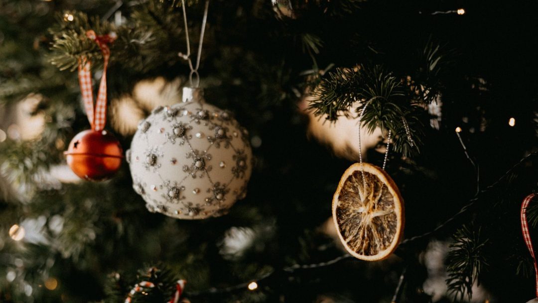 Julentreet glitrer med ornamenter, inkludert en rød klokke, en hvit kule utsmykket med sølvmønstre og en tørket appelsinskive.