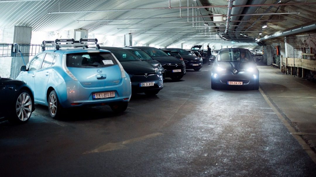 I et svakt opplyst underjordisk parkeringsområde går flere biler på tomgang til venstre mens en med frontlykter på innkjøring fra høyre, med tanke på Klimagassregnskapets standarder. Takrør veves over, og legger til den urbane atmosfæren.