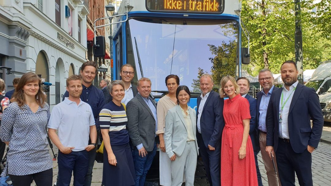 En gruppe på tolv personer som står foran en stillestående blå buss i en brosteinsgate omkranset av bygninger og trær. Bussen viser «Ikke i trafikk.