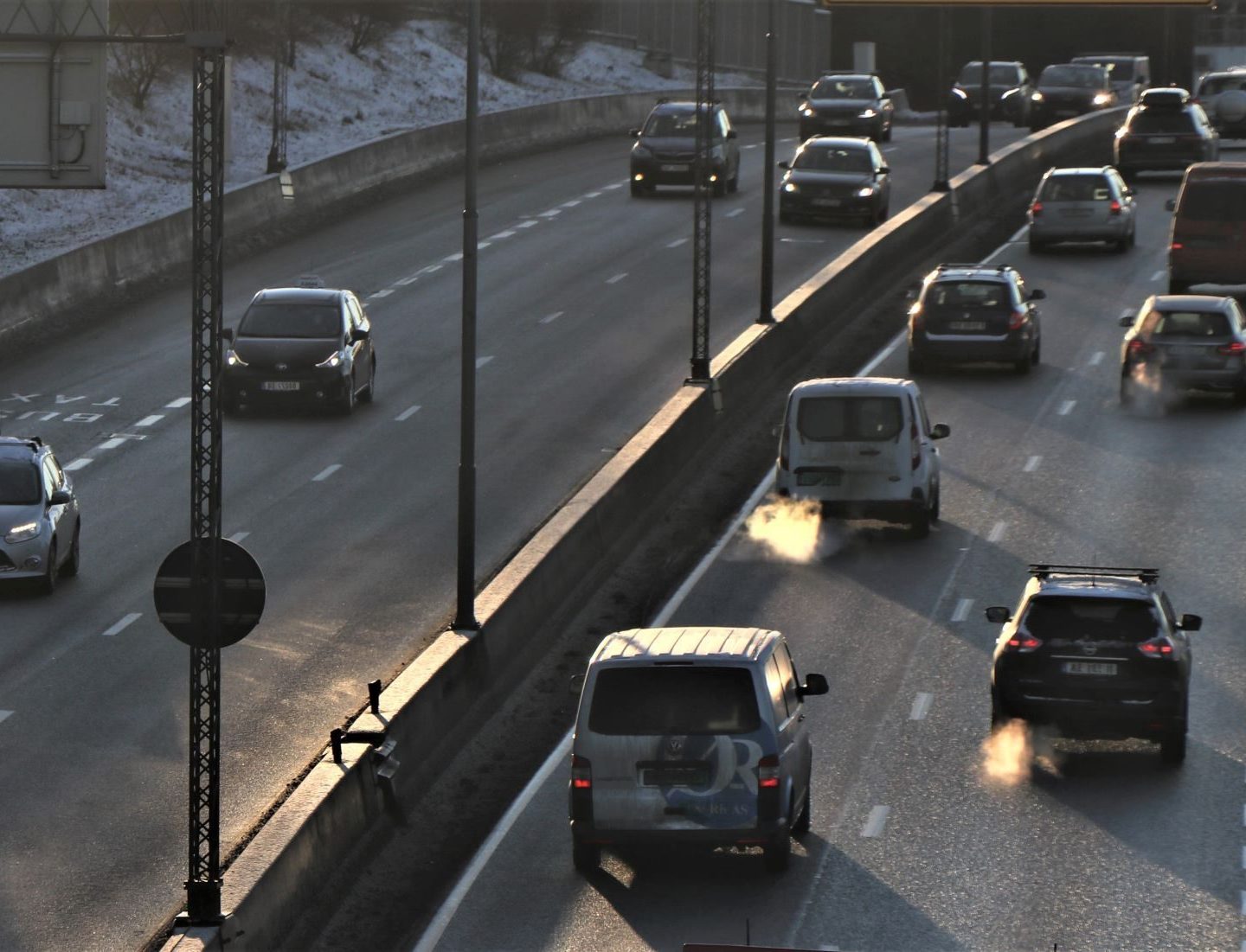 Biler som kjører på en flerfelts motorvei i dagslys, med noen kjøretøyer som slipper ut synlig eksos. Trafikken fremstår som moderat, og det er skygger på veien.