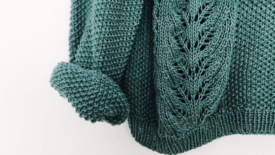 Et nærbilde av en grønn strikket genser, som viser et julen-inspirert blondemønster og opprullede ermer, satt mot en ensfarget hvit bakgrunn.