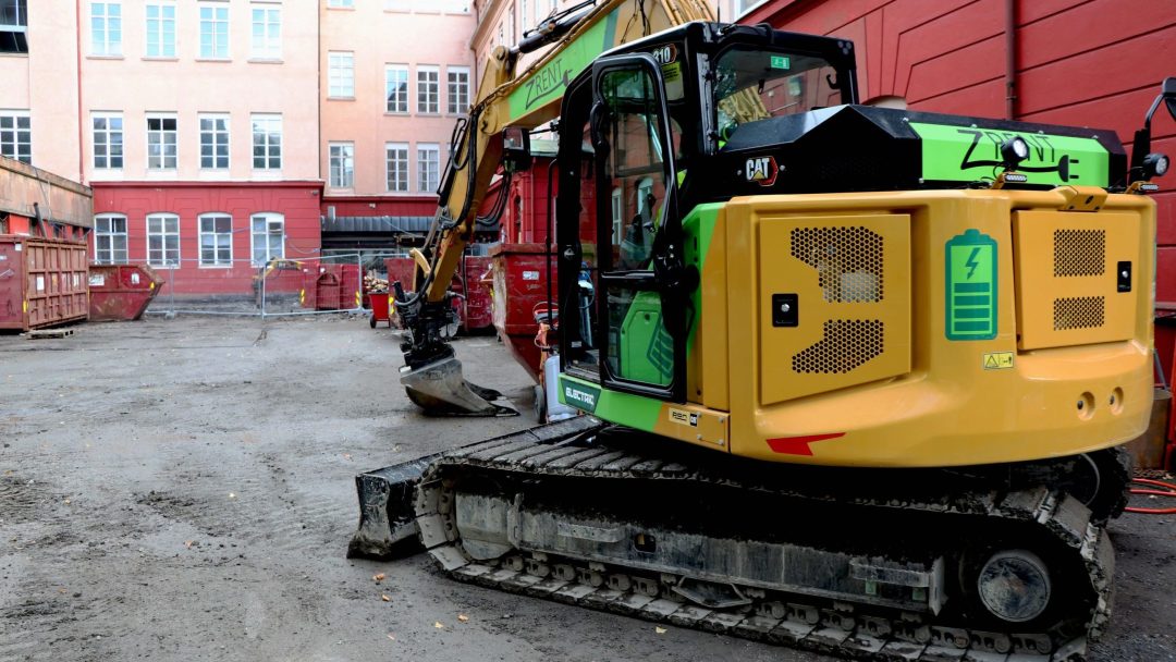 En grønn og gul gravemaskin er parkert indirekte ved siden av en byggeplass, innrammet av de livlige fargetonene til røde og rosa bygninger i bakgrunnen.
