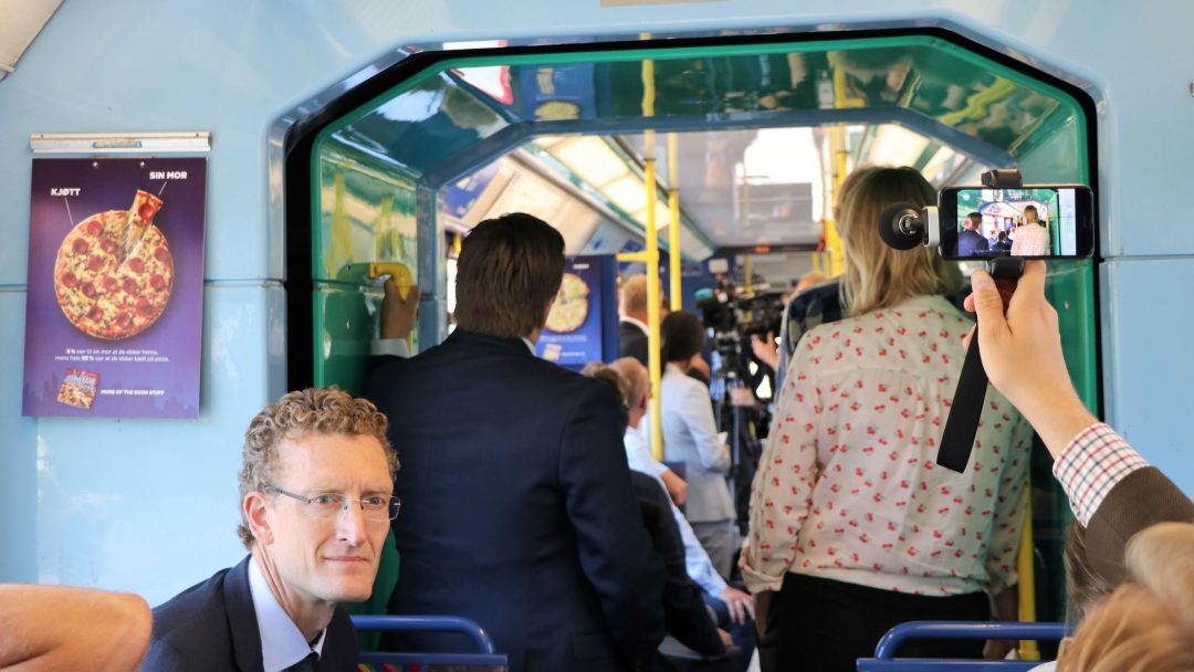 Folk som står og sitter inne i en trikk, noen ser på et kamera som tar opp scenen. Plakater og gule stolper er synlige, og en person holder opp en smarttelefon.