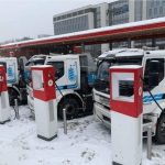 Elektriske lastebiler parkert ved ladestasjoner i et snødekt område nær en moderne bygning.